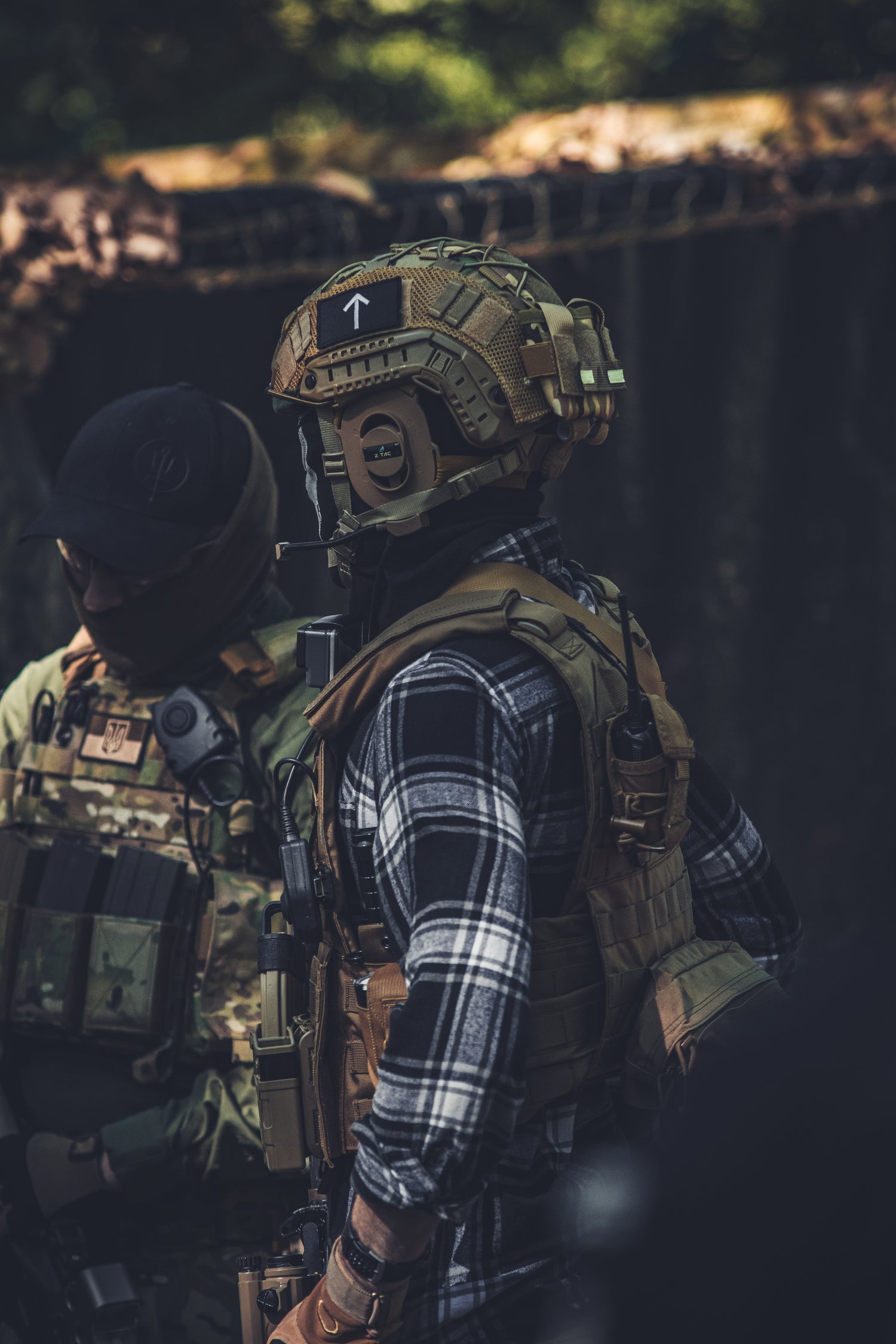 A person in tactical gear stands outdoors wearing a tan helmet with a mounted night vision device, a black-and-white plaid flannel shirt, and a tan tactical vest. Another person in camouflage gear is partially visible beside them in a dark, wooded setting.