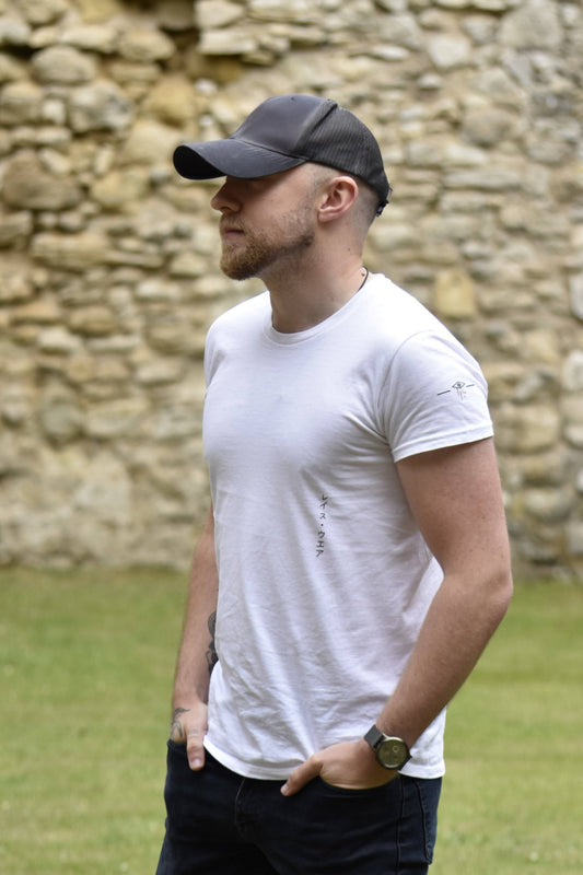 Man wearing a white t-shirt and dark cap standing in front of a stone wall.