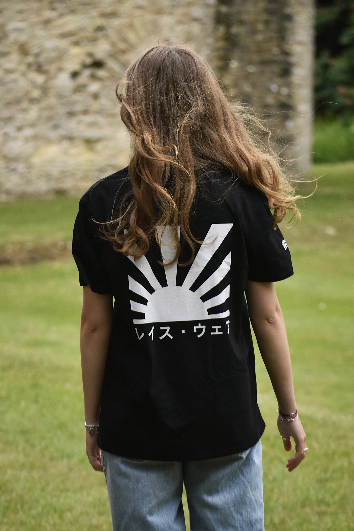 Person wearing a black t-shirt with a white graphic design, standing in a grassy area with a stone wall in the background.
