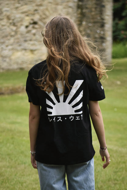 Person wearing a black t-shirt with a white graphic design, standing in a grassy area with a stone wall in the background.