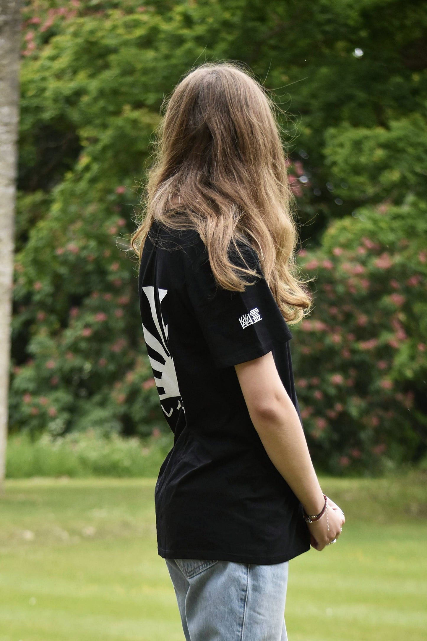 Person wearing a black t-shirt with a logo and light blue jeans standing in a grassy area with trees and ruins in the background.